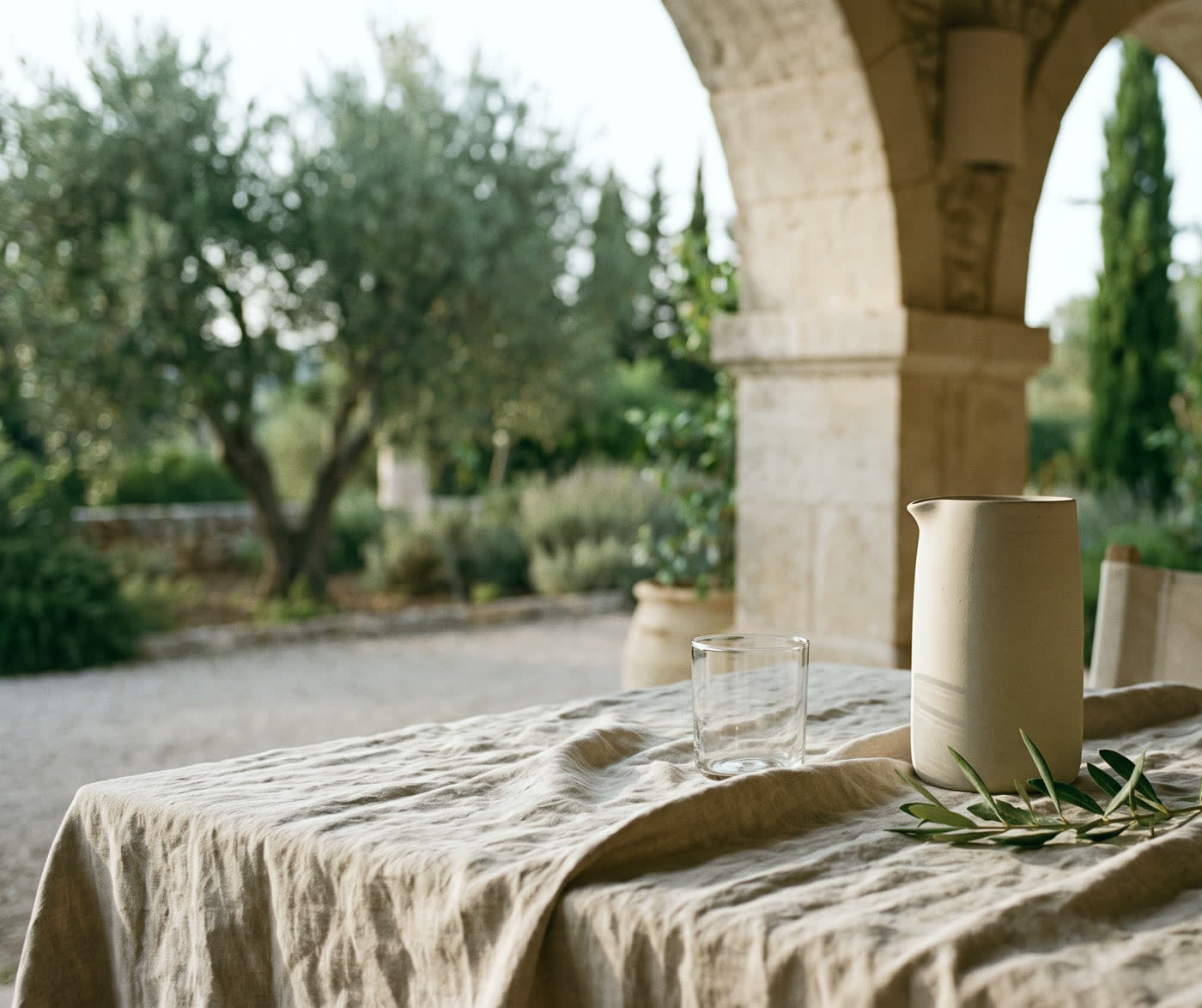 A linen-covered table in a shaded stone loggia, set with a ceramic carafe, a glass and a sprig of olive.